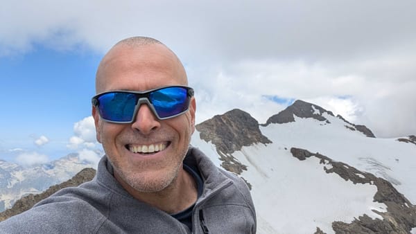 My smiling face, in blue sunglasses, on a summit with a glacier and snowy peaks behind me